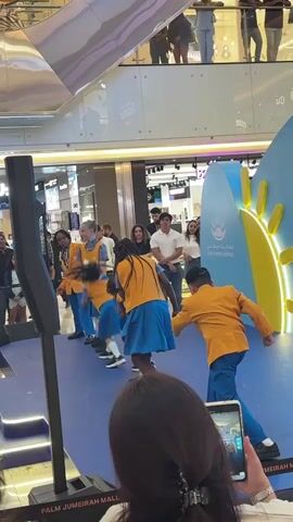 The video shows a group of children in matching orange shirts and blue skirts or shorts performing a synchronized dance routine on a blue stage in a shopping mall. The children are lined up in a row, and one child in the back right is moving their arm up and down. The stage is set up in front of a large blue and yellow backdrop with palm tree graphics and Arabic text. There are spectators standing around the stage, many of whom are filming the performance with their phones. In the foreground, a person is holding a phone and recording the event. A tall black speaker is positioned to the left of the stage. The overall environment suggests a public event or performance taking place in a commercial space.