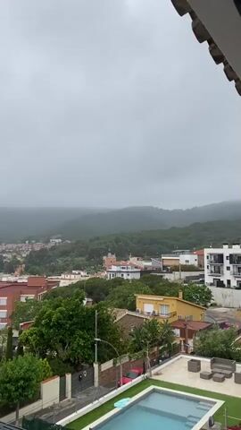 The video captures an elevated, panoramic view of a residential area in Vilassar De Dalt, Spain, on an overcast day. The sky is completely gray, and the distant hills and mountains are shrouded in mist or low clouds, suggesting a cloudy or foggy weather condition.

In the foreground, a swimming pool with a blue-green hue is visible on the right, surrounded by paved areas and some greenery. Adjacent to the pool are modern outdoor furniture pieces. The camera appears to be positioned on a balcony, with a glimpse of a terracotta-tiled roof edge in the top right.

The midground features a cluster of houses and buildings with varying architectural styles, many featuring red or terracotta tile roofs. Colors range from warm yellows and browns to white and some with reddish-brick facades. Lush green trees and vegetation are abundant throughout the scene, interspersed between the structures and along the streets. A street with parked cars can be seen winding through the residential area.

In the background, densely forested hills rise, partially obscured by the mist, creating a soft, ethereal quality. A small, white object, which appears to be a drone, briefly flies across the frame in the early part of the video (around 0:01), accompanied by an audible buzzing sound throughout, indicating its proximity. The camera slowly pans from right to left, revealing more of the town and the surrounding natural landscape. The overall atmosphere is calm and subdued, reflecting the cloudy conditions.