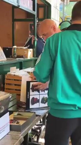A bald man in a black shirt carefully examines a Gorillaz record at an outdoor vendor stall. He is standing in front of a table laden with various items, including wooden crates filled with records. Another man, wearing a green Adidas track jacket, stands next to him, also looking at the record. In the background, a person wearing an Adidas tracksuit and another with a green cap are visible. The vendor stall is constructed from wood and has several shelves displaying merchandise. The setting seems to be a busy market or fair.