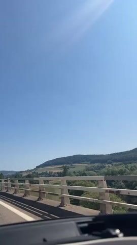 The video shows a view from a car driving over a bridge on a sunny day. The sky is clear and blue with the sun creating a lens flare in the upper right corner. The bridge has white railings and a concrete barrier. The landscape visible on either side of the bridge consists of rolling green hills and fields, with scattered trees and some buildings in the distance. Several cars and trucks are seen on the road, including a white RV. The camera appears to be mounted inside the car, looking forward.