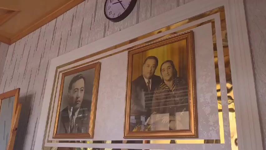 A slow-motion, medium shot from a low angle shows two framed portraits on a wall. On the left, a man in a suit and tie is captured in a black and white photograph. To the right of this portrait, a color photograph features a man and a woman, both dressed formally, posing together. The portraits are mounted within a larger, decorative frame that incorporates mirrored elements. Above the portraits, a wall clock with a white face and black hands is visible. The wall itself is covered with striped wallpaper. To the left of the portraits, a freestanding mirror with a wooden easel is partially visible. The lighting appears to be natural and warm.