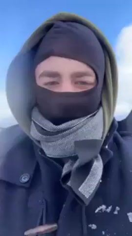 The video shows a person wearing a black balaclava, grey scarf, green hoodie, and dark blue jacket, looking directly at the camera. They are outside on a bright day, likely in a cold environment. The person says, "So, guys, this is the second day exploring". Another person, mostly obscured by the first person's arm, briefly appears behind them. The background features a bright, cloudy sky and a snow-covered landscape.
