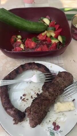 Here's a brief description of the video:

The video shows a person's hand dipping a piece of bread into a salad in a burgundy bowl. The salad appears to have tomatoes, cucumbers, and green onions. Next to the bowl is a plate with two grilled sausages and two forks. A can and a salt shaker are visible in the background on a beige tablecloth. The speaker in the video says, "In Bulgaria, we eat like this."