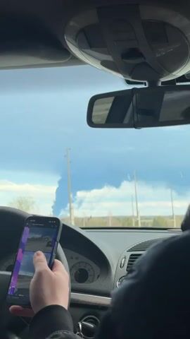 From inside a car, a plume of dark smoke is visible in the distance on a bright, sunny day. The landscape consists of a road, power lines, and green fields or forest. A person in the passenger seat is holding a phone, appearing to watch a video on it that depicts the same smoke plume. The interior of the car is visible, including the dashboard, steering wheel, and rearview mirror. A person is driving.