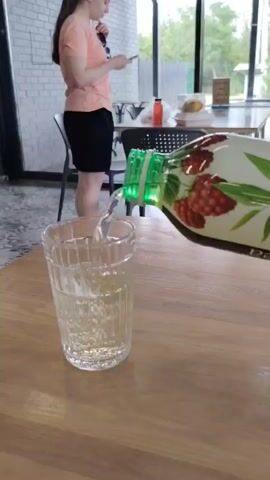 A person wearing a peach t-shirt and black shorts is standing in the background. In the foreground, a bottle with a green cap is pouring a yellow liquid into a clear glass. The liquid creates bubbles as it fills the glass. Another glass is visible behind it. The scene takes place at a wooden table.