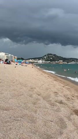 The video shows a sandy beach with several people swimming in the turquoise water. The sky is overcast with dark, ominous clouds, suggesting an approaching storm. Buildings are visible in the background on a hillside overlooking the beach. Some people are sitting on the sand, and others are walking along the shoreline. Waves are gently lapping at the shore. The overall scene is a beach with people enjoying themselves despite the impending weather.