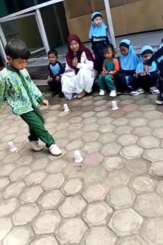 A group of young children, some wearing hijabs and blue headscarves, are seated outdoors on a paved patio area with a hexagonal tile pattern. A boy in a green and white patterned shirt and green pants is actively engaged in a game, kicking at small white cups scattered on the ground. The boy is in mid-action, his body angled as he appears to be making a kick or movement. Several other children, and an adult woman in a hijab, watch him. The environment appears to be a school or community setting, with a building and some greenery visible in the background.