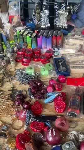 The video shows a market stall laden with various items. The foreground is dominated by a cluttered wooden table displaying an assortment of keychains, many of which are shaped like pomegranates and some are red or pink. There are also numerous small knives and lighters in a variety of colors, including green, pink, and blue. Scattered amongst these are some beaded necklaces and bracelets in assorted colors. In the background, more shelves are visible, filled with more rows of lighters, small bottles, decorative items such as figurines and wall plates, and stacks of boxes. The overall impression is of a busy, diverse market stall selling a wide range of trinkets and accessories.