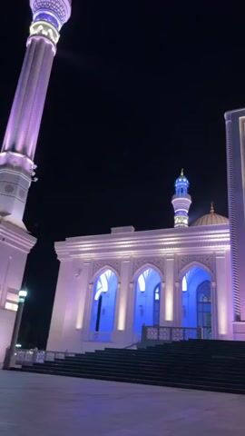 The video shows a mosque at night. The mosque is illuminated with purple and blue lights. There is a minaret on the left side of the mosque, and another minaret on the right side. In front of the mosque, there are steps leading up to the entrance. There is also a fountain in front of the mosque that is lit with blue and pink lights. The sky is dark and clear.