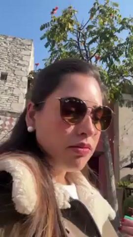 A woman with long brown hair is seen from the side, wearing sunglasses, a white shirt, and a cream-colored jacket. She is outdoors on a sunny day, with a tree to her right and a stone building in the background. There's a red building partially visible behind the tree. The camera seems to be hand-held, as it shifts slightly.