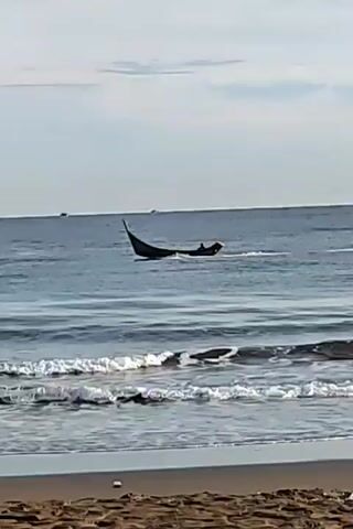 A small, dark boat with a pointed bow is moving across choppy, blue ocean water. The boat appears to have at least one person inside. The waves are breaking onto a sandy beach in the foreground. The sky is overcast and pale.