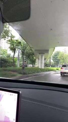 The video begins from the interior of a vehicle, looking out through the windshield. The windshield is wet with raindrops and streaky. Outside, a car is driving on a wet road under an elevated structure. The road appears to be a multi-lane road with a grassy median and trees lining the sides. In the distance, several tall buildings can be seen. A white SUV is visible ahead, driving in the same direction as the camera's vehicle. A blue road sign with white text and arrows is present on the right side of the road. The elevated structure has large concrete pillars supporting it. The overall environment suggests an urban setting during a rainy day. The vehicle's dashboard display is partially visible on the bottom left of the screen.