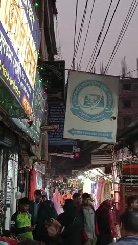 The video shows a narrow, bustling street in Bangladesh. Numerous shops line the street, their signs written in Bengali. Colorful lights are strung across the street, adding to the visual clutter. A large crowd of people, mostly men, move through the narrow space. Overhead, a dense network of electrical wires create a chaotic web against the overcast sky. The overall impression is one of a busy and crowded commercial area.