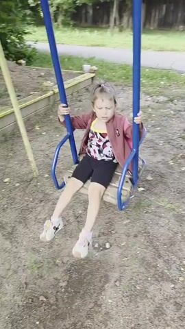 A young child with blonde hair tied in a ponytail is on a blue metal swing set in a sandy, grassy area. The child is wearing a maroon jacket over a white and red patterned shirt and black shorts. The child is holding onto the blue bars of the swing and is gently swaying back and forth. In the background, there is a wooden fence and some trees. The lighting suggests it is daytime.