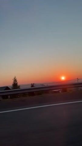 The video begins by showing a scenic view of a sunset over a rural landscape with houses and trees visible in the distance. A highway lane marked with white lines is in the foreground. A red SUV drives by in the second frame, with a person visible in the driver's seat. The subsequent frames continue to show the sunset and the highway, with no further action or interaction.