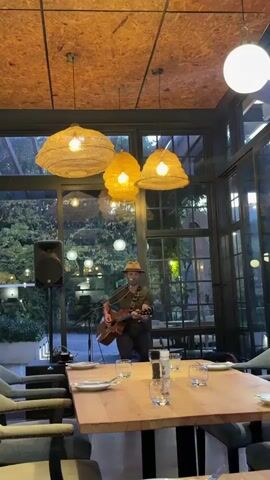 Here is a description of the video. 

The video shows a man playing an acoustic guitar and singing into a microphone in a restaurant. He is wearing a hat and a dark green shirt and pants. He stands behind a long, empty wooden table that is set with plates, glasses, and salt and pepper shakers. A black speaker stands to his left. The restaurant has large windows looking out onto a courtyard with greenery and hanging lights. Above the table are three large, woven pendant lamps. The ceiling has an exposed plywood-like surface.
