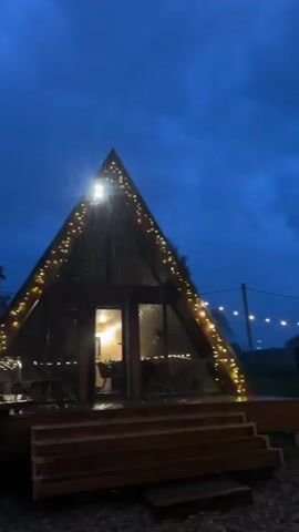 The video opens with a view of an A-frame cabin, illuminated by string lights, against a backdrop of a dark blue evening sky with rain. The camera pans left to reveal a wooden hot tub next to the cabin, with a tall, thin chimney rising from it. The final shot shows a low-angle view of the chimney and the hot tub's edge, with neighborhood houses and utility poles visible in the background.