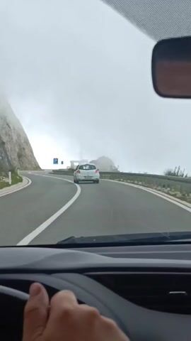 The video, filmed from the driver's perspective inside a car, shows a winding mountain road on a foggy day. The driver's hand is visible on the steering wheel. A white car is ahead, also driving on the road. To the left, a steep, rocky cliff face is covered in fog. To the right, a guardrail lines the side of the road, and a blue parking sign is visible. The sky is overcast and the visibility is limited by the fog.