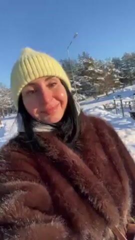 A woman is holding the camera and recording a video of herself in a snowy park. She is wearing a brown fur coat and a yellow knitted hat. The sky is clear and blue. There are trees covered in snow in the background. The woman appears to be smiling slightly at the camera.