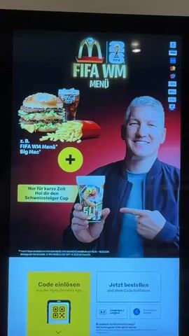 The video shows a digital menu screen at a McDonald's restaurant. The screen displays a promotion for a FIFA World Cup menu, featuring a Big Mac, fries, and a drink. A man, identified as Bastian Schweinsteiger, is shown holding a promotional cup and pointing towards it. The bottom of the screen shows options to redeem a code through the McDonald's app or to proceed with an order without a code. The video pans to a close-up of the Big Mac. The menu is in German.