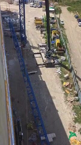 This is a high-angle aerial view of a construction site. A blue tower crane is visible on the left, extending vertically. At the top of the tower crane, a person wearing orange and a hard hat is visible, working near the hook mechanism. On the right side of the frame, a large mobile crane is positioned with its outriggers extended for stability. The mobile crane is predominantly green and black, with a visible operator's cab. The ground is a mix of gravel and dirt, with various construction materials scattered around, including stacks of yellow planks, concrete blocks, and wooden debris. A dirt path runs between the tower crane and the mobile crane. In the background, a white building or structure and a few parked vehicles are discernible. The lighting suggests it is daytime with sunlight casting shadows.