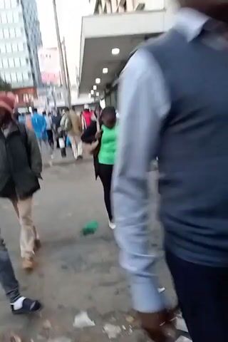 The video shows a street scene, possibly a market area, with numerous people walking around. The ground is littered with trash. Several people are wearing casual clothing, including a woman in a headscarf, a man in a jacket with a backpack, and a woman in a green shirt. A man is visible talking on a cell phone. There is a small food cart with a glass display case. The background shows buildings with closed metal shutters. The scene appears busy and urban.