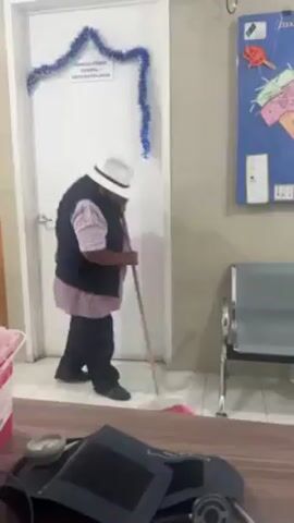 The video shows a person sweeping a floor with a broom in what appears to be a medical or dental waiting room.

A person, wearing a white hat, a dark vest, and dark pants, is sweeping the floor with a long-handled broom. They sweep near a door, which has a blue garland above it and a sign on it that reads "CONSULTORIO DENTAL Y ESTOMATOLOGIA." The person is facing the door.

To the right of the door are three metal chairs, and a blue map or chart is hanging on the wall above the chairs. A red biohazard container is partially visible in the lower left of the frame, along with a sphygmomanometer on a desk. The floor is tiled.