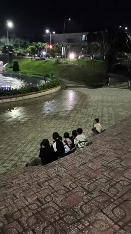At night, a group of six people, likely children, sit on a set of tiled steps facing a large, round fountain. The fountain shoots jets of water lit up by various colors, creating a dynamic light show. In the background, city lights twinkle, and buildings can be seen. The atmosphere is relaxed and possibly rainy, as the tiled surface looks wet and reflective.