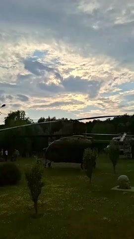 The video opens with a wide shot of an outdoor display area under a cloudy sky. In the foreground, a green lawn is dotted with small trees and bushes. A gravel path leads into the scene. Various objects are scattered across the lawn, including what appear to be sculptures and some large structures resembling aircraft. The first prominent object is a large, dark green helicopter-like structure with its blades visible. To the right of the helicopter, a smaller, white structure resembling an airplane with visible propellers is present. Further into the scene, more sculptures and what looks like another airplane are visible. The overall impression is of an open-air exhibition or park, possibly featuring decommissioned aircraft and artistic installations. The lighting suggests it might be late afternoon or early evening. The video then pans across the landscape, showcasing more of the green expanse and the scattered exhibits, including several human-like statues or busts. The environment is natural with trees forming a backdrop. There are no people visible interacting with the exhibits.
