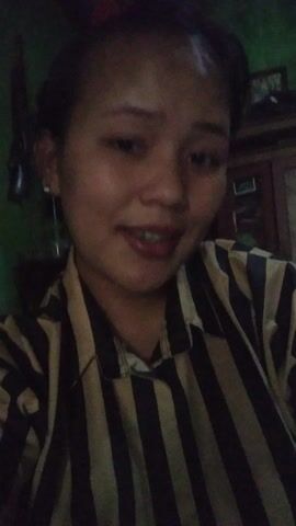 A woman is featured in a close-up video, angled sideways. She has dark hair and is wearing a striped shirt with black and yellow or beige stripes. She is looking directly at the camera, smiling, and appears to be speaking or singing. Her mouth is open in some frames, showing her teeth. The lighting is dim, suggesting it might be evening or night. The background is dark and indistinct. An audio overlay features a woman's voice speaking or singing in Indonesian, with phrases like "Kamu, kok seperti hantu..." (You, why are you like a ghost...). The scene appears to be indoors in Kecamatan Cimaung, Indonesia.