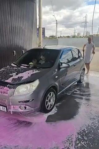 The video begins with a close-up of pink foam and water gushing onto a wet pavement. A car wheel, partially visible on the left, is covered in foam. The camera then pans up to reveal a person standing under a canopy. The person is wearing a white t-shirt and dark shorts. The canopy appears to be made of white fabric stretched over a metal frame. The person is looking up, possibly at the sky or the canopy itself. The scene then cuts to a close-up of the white fabric of the canopy. Finally, the camera is looking down at the feet of a person wearing black shorts and black and white sneakers, standing in pink foam and water on the pavement. The person is looking down at the foam.