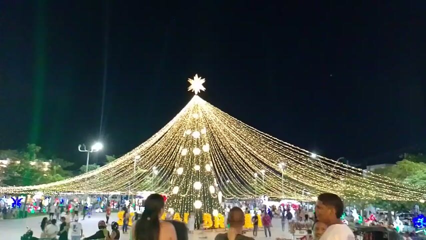 The video depicts a night scene in what appears to be a town square or public gathering space. The focal point is a large, illuminated structure resembling a Christmas tree, decorated with numerous strings of lights culminating in a star at the top. Several people are walking around the area near the base of the "tree", suggesting pedestrian activity. Street lights are visible and there is a hint of buildings in the periphery, contributing to the overall ambience. The sky is dark, adding to the nighttime setting.