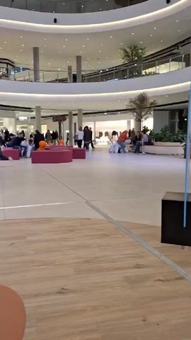 The video shows the interior of a large, modern shopping mall. The mall has multiple levels, with open walkways overlooking the main floor. There are several stores visible along the walkways, including Levi's and Oysho. The main floor is spacious, with seating areas featuring bright pink cylindrical benches and orange, dog-shaped ride-on toys. People are walking around, sitting on the benches, or shopping. A child in a blue shirt and another in an orange shirt are riding the dog-shaped toys. The flooring of the main area is light beige tile, while the area in front of the camera is wood-grained. The lighting is bright, and the overall impression is of a clean and modern shopping environment.