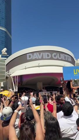 A large crowd of people stand with their arms raised, each holding up a smartphone, likely recording a performance. In the background, a building has "DAVID GUETTA" displayed in large white letters on a digital screen. A blue sign with "LIV BEACH" printed on it is held to the side. The sky is a clear blue. The scene suggests an outdoor concert or music event featuring David Guetta.