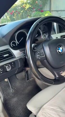 The video pans across the interior of a BMW car. The focus is on the steering wheel, dashboard, and the passenger seat. A plush toy bunny wearing a red sweater with the BMW logo is visible on the passenger seat. The exterior environment, including greenery and a fence, is visible through the car windows.