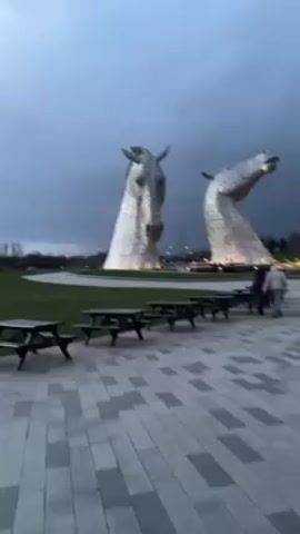 The video shows two large, silver-colored horse head sculptures standing in a grassy field. The sky is overcast with dark clouds. In the foreground, there is a paved area with several black picnic tables. Three people are standing near the picnic tables, looking towards the horse heads. There are trees visible in the background, along with utility poles. The lighting suggests it is dusk or early evening.