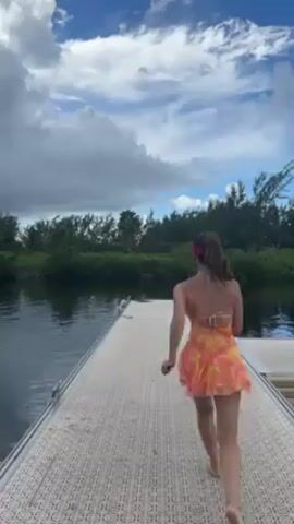 A woman in an orange, yellow, and white dress walks to the end of a pier. She then turns around, pauses, and then continues walking forward. The pier is constructed of interlocking gray tiles. On either side of the pier is dark, calm water. A green, lush shoreline is visible beyond the water. The sky is blue with large, fluffy white clouds.