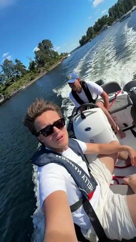 The video shows two young men on a rigid inflatable boat on a body of water. The man in the foreground is wearing sunglasses and a white t-shirt, and is holding a phone up as if taking a selfie. He is sitting on the left side of the boat. The second man, wearing a white t-shirt and a blue cap, is seated behind the first man and appears to be steering the boat. Both men are wearing life jackets. The boat is moving at a moderate speed, leaving a wake behind it. The background shows a shoreline with trees and houses, and several sailboats moored in the distance. The sky is blue with some scattered clouds, and the sun is shining.