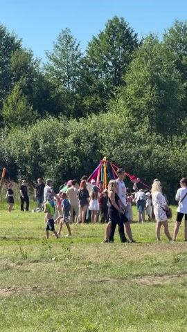 The video shows a group of people gathered in a grassy field on a sunny day, with trees in the background. In the center of the gathering is a tall pole adorned with colorful ribbons, suggesting a festive or traditional event. Many of the people appear to be women and children, some of whom are dressed in white or light-colored clothing. There are various interactions happening: some people are standing and observing, while others, particularly children, are moving around the field. A few couples are visible on the right side of the frame. The overall atmosphere seems relaxed and celebratory.
