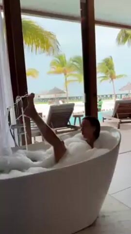 A woman is relaxing in a white, freestanding bathtub filled with bubbles, with her leg sticking out. She appears to be in a bright, airy room with large windows showcasing a tropical beach scene with palm trees, lounge chairs, and thatched umbrellas. The woman is smiling and seems to be enjoying herself. A chrome faucet is running into the tub. The background shows a wooden deck leading to the beach and the overcast sky suggests a cloudy day.