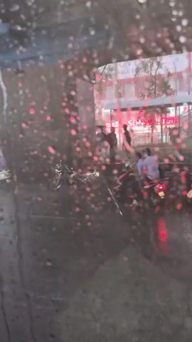 The video shows raindrops on a window, blurring the view of a street scene. There are cars and possibly motorcycles moving in the distance, with red lights visible. It appears to be nighttime or a very overcast day, given the artificial lighting. The overall impression is of a rainy urban environment viewed from inside a vehicle.