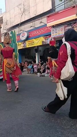 A parade is underway on a street. In the foreground, a woman in a black hijab and maroon jacket is holding white flip-flops. Behind her, a decorated float is moving down the street. On the float, there are people in traditional costumes. Several children and adults are watching the parade from the side of the road. The buildings lining the street appear to be shops. The sky is overcast.
