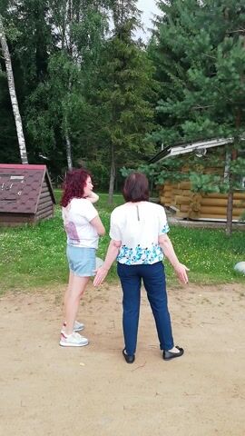 In this video, two women are outdoors in a sandy area surrounded by trees and grass. The scene appears to be rural, possibly a park or a backyard. The woman in the foreground, with curly red hair, wears a white t-shirt and denim shorts. The woman behind her is wearing a white blouse with a floral pattern and dark pants. Both women are facing away from the camera, with their arms outstretched. The lighting suggests it is daytime, with overcast or diffused sunlight. There is a small wooden structure visible in the background. The overall mood seems casual and relaxed, perhaps indicative of a recreational activity or a simple outdoor gathering. The video is oriented vertically, suggesting it was filmed with a mobile phone.