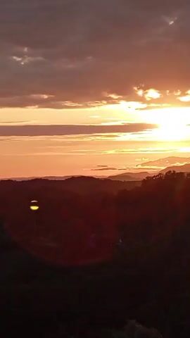 The video shows a sunset over a hilly landscape. The sun is setting in the background, casting a warm orange glow across the sky. Clouds are visible in the sky, with some illuminated by the sun. In the foreground, there are dark silhouettes of hills and trees. A circular lens flare is prominent in the lower portion of the frame.
