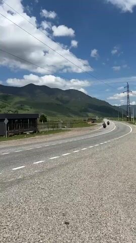 A vertical video shot from the perspective of someone moving along a paved road, likely on a motorcycle or in a vehicle. The road has white dashed lines marking the lanes. To the left of the road, there are several dark, modern-looking buildings with wooden accents and some fencing. Beyond these buildings, there are green, rolling hills that transition into larger mountains in the background. The sky is blue with scattered white clouds. A few motorcycles are visible on the road, moving in the same direction as the camera. Further down the road, there are more buildings, some vehicles, and what appears to be construction equipment. The scene suggests a rural or mountainous landscape with some infrastructure.