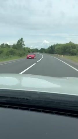 A car is driving on a highway. The road is empty except for one other car far ahead. The sky is cloudy and the trees on both sides of the road are green.