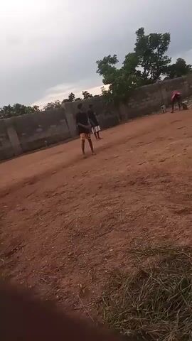 The video shows a dirt field with a brick wall in the background. Several people are on the field, and they appear to be playing soccer. The camera is shaking slightly. The weather is overcast.