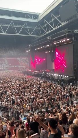 The video captures a vibrant concert scene inside a large stadium, packed with a massive crowd of enthusiastic attendees. The stage is illuminated with bright pink and red lights, featuring large screens displaying abstract visuals. A significant sound system is suspended above the stage. The audience, a dense sea of people, are actively engaged, with many holding up their phones to record the event. The atmosphere appears energetic and celebratory. The time of day seems to be daytime, judging by the natural light filtering in from the open roof of the stadium, although the interior is dominated by artificial stage lighting. Visible text in the background reads "DÜSSELDORF ES IST EIN GROSSARTIGER TAG," which translates to "Dusseldorf, it is a great day," providing a clear location cue. The overall impression is one of a lively and memorable musical event.