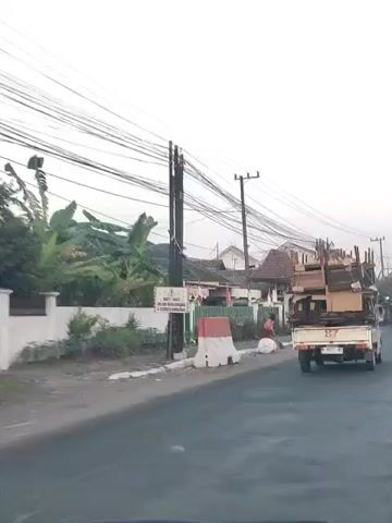 The video shows a street scene with a white pickup truck loaded with furniture, possibly being transported for disposal or reuse. A person is sweeping the sidewalk near a large white cylindrical object and a signpost. The truck has a license plate that reads "B 8671 WE". Houses decorated with Indonesian flags are visible in the background, suggesting a patriotic or festive occasion. Overhead, a dense network of electrical wires crisscrosses the sky. The overall impression is that of a daily life scene in a residential area, possibly during a time of cleanup or preparation for an event.