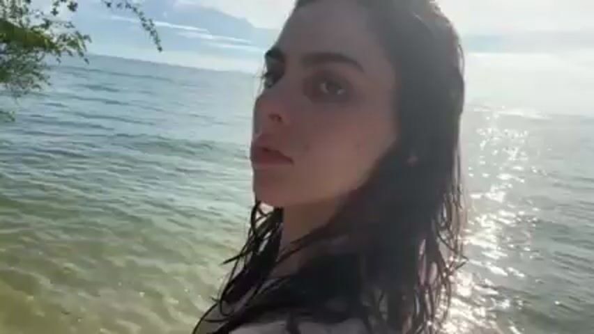 A woman in a black bikini is standing in shallow water on a beach. The water is clear and the sand is light-colored. The woman is looking at the camera. Her hair is dark and wet. There are small pieces of seaweed or debris floating in the water. The camera angle is from a low perspective, looking up at the woman. She is moving through the water, causing ripples. The scene is bright and sunny.